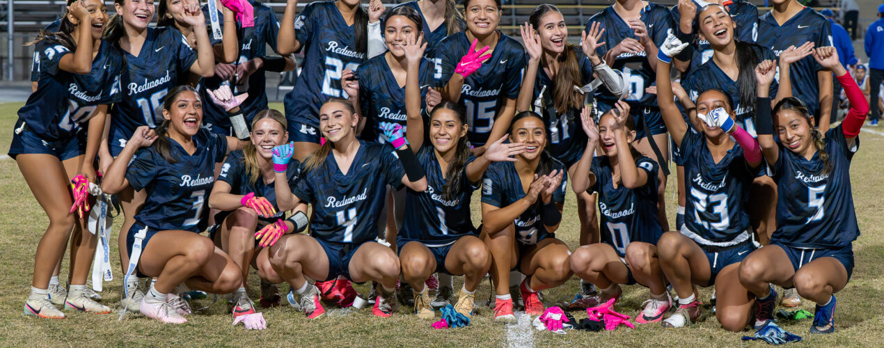 Redwood players crouch and stand together on the field for a group photo, cheering, waving and smiling.