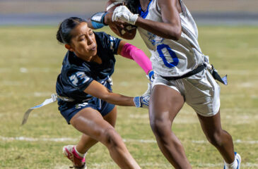 A Madera ball carrier holding the football runs upfield while a Redwood defender lunges toward her waist to grab the flag on her belt.