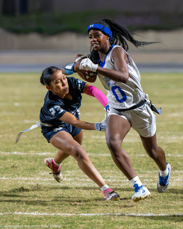 A Madera ball carrier holding the football runs upfield while a Redwood defender lunges toward her waist to grab the flag on her belt.